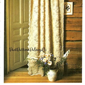 Op de afbeelding: Een gehaakt gordijn met een geometrisch patroon hangt in een rustieke houten kamer. Het gordijn is crèmekleurig en heeft een gegolfde zoom. Een vaas met bloemen staat op een houten oppervlak voor het gordijn.