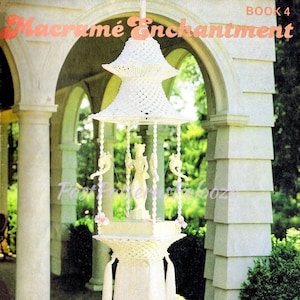 May include: A white macrame hanging sculpture, resembling a tiered pagoda, with decorative figures and tassels. The sculpture is suspended from a hook and features the text "Macrame Enchantment" and "Fountain of the Orient".