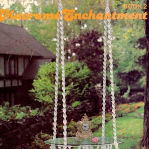 以下が含まれることがあります： 白いマクラメのテーブルで、ガラスの天板とタッセルが下からぶら下がっています。テーブルは4本の白いコードで天井から吊り下げられています。テーブルの上には時計とピンクの花があります。画像の上部には「Macrame Enchantment Book 2」というテキストがあります。画像の下部には「Fantasy」というテキストがあります。