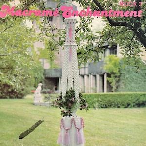 Puede incluir: Un macetero de macramé blanco con borlas rosas y una cinta rosa. El macetero está colgado de una rama de árbol en un jardín. El título "Macrame Enchantment Book 3" y "Country Lass" son visibles en la imagen.