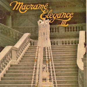 以下が含まれることがあります： 白いマクラメのウェディングケーキスタンドに、白いウェディングケーキが入っています。スタンドは、白いマクラメの天井照明器具から吊り下げられています。スタンドは、白い大理石の階段の前にあります。画像は、「マクラメ エレガンス II」というタイトルの本からのものです。