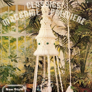 May include: A white macrame plant hanger with a green beaded trim. The hanger is suspended from the ceiling and has a round base. The hanger is in a greenhouse setting with plants in the background.