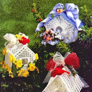 Op de afbeelding: Drie gehaakte vogelhuisjes versierd met linten en kunstbloemen. De vogelhuisjes zijn wit met blauwe, gele en rode accenten. Elk vogelhuisje heeft een klein vogeltje erin.