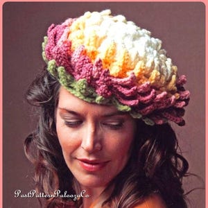 May include: A woman wearing a brown leather jacket with a crocheted flower hat in shades of pink, yellow, and white. The hat is a multi-layered design with a green crocheted base.