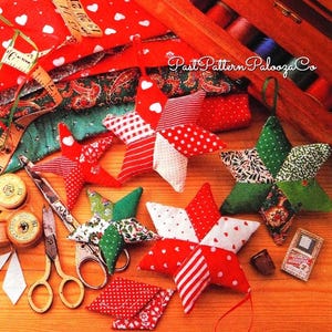 May include: A collection of handmade Christmas star ornaments in red, white, and green patchwork fabrics. The stars are arranged on a wooden surface with sewing supplies, including scissors, spools of thread, and fabric scraps. The image includes the text "Past Pattern Palooza Co."