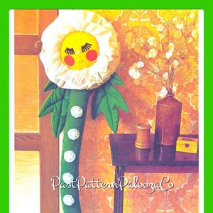 Puede incluir: Una flor de tela blanca con una cara amarilla y mejillas rojas. La flor está unida a un tallo verde con botones blancos. La flor está apoyada contra una pared con papel tapiz floral naranja y marrón.