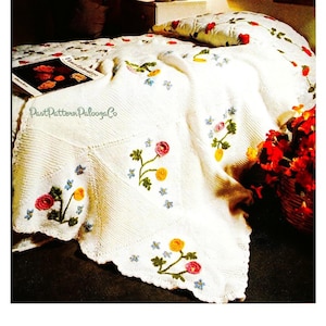 Puede incluir: Manta de crochet blanca con borde festoneado y diseños florales bordados en tonos rosa, amarillo y azul. La manta se muestra en una cama con una almohada con estampado floral.