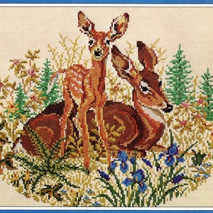 May include: Cross-stitch embroidery of two brown deer in a forest setting. The deer are surrounded by green trees, blue flowers, and yellow wildflowers.