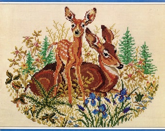 May include: Cross-stitch embroidery of two brown deer in a forest setting. The deer are surrounded by green trees, blue flowers, and yellow wildflowers.