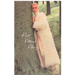May include: A woman wearing a long, white, shaggy knit cardigan with a turtleneck sweater underneath. She is standing in a forest with a tree behind her.