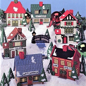 May include: A set of seven miniature Christmas village houses made of plastic canvas. The houses are decorated with red, green and white paint and feature various shop names, including "Fish Shoppe", "Gift Shoppe", "Bakery", "Tailor", "Watchmaker", and "Wine Spirits".