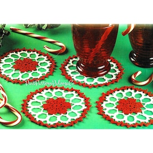 May include: Four handmade crochet coasters with a red and white snowflake design are displayed on a green surface. The image includes candy canes and two glasses of a red beverage, evoking a festive atmosphere.