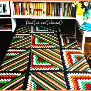 May include: A colorful, handmade crochet blanket with a geometric pattern. The blanket features squares with diagonal stripes in red, green, orange, white, and black. The blanket is displayed on a bed, with a bookshelf and other items in the background.