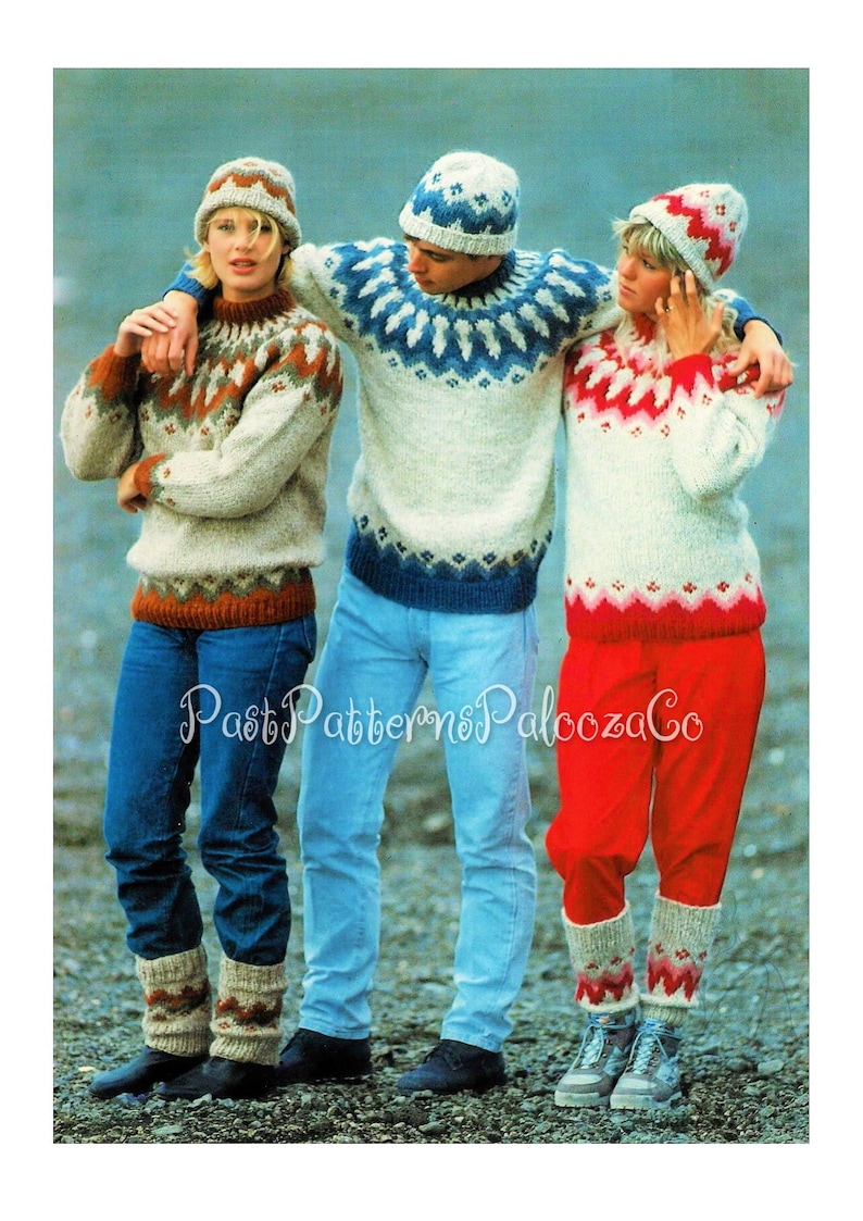 May include: Three people wearing knitted sweaters and hats with traditional Norwegian patterns. The sweaters are white with blue, red, and brown accents. The hats are white with blue and red accents. The people are standing on a rocky beach.
