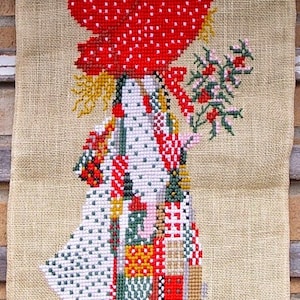 May include: A cross-stitch wall hanging featuring a girl in a red and white polka dot hat and a patchwork dress holding a sprig of red berries. The girl is standing on a burlap background.