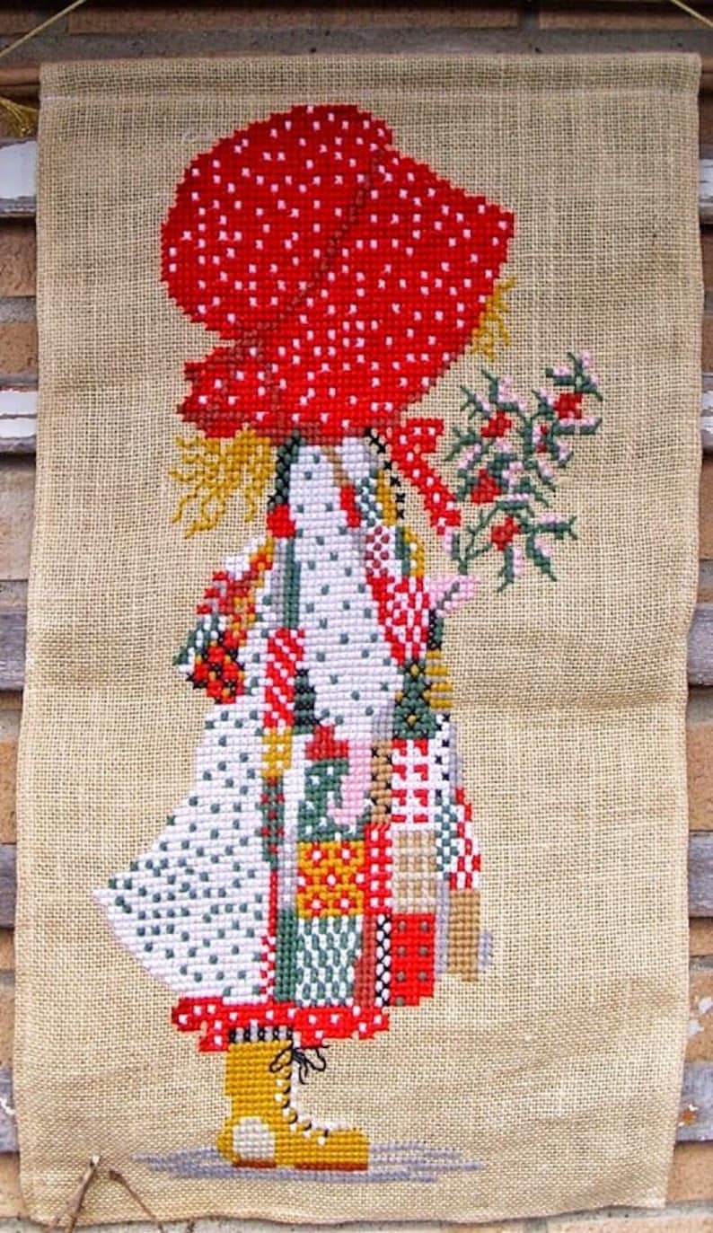 May include: A cross-stitch wall hanging featuring a girl in a red and white polka dot hat and a patchwork dress holding a sprig of red berries. The girl is standing on a burlap background.