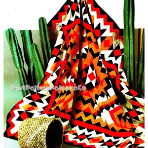 Pode incluir: Uma colcha geométrica vermelha, laranja, preta e branca com uma borda em ziguezague. A colcha está dobrada e drapeada sobre uma cesta tecida. O fundo é uma superfície branca com plantas de cacto verdes.