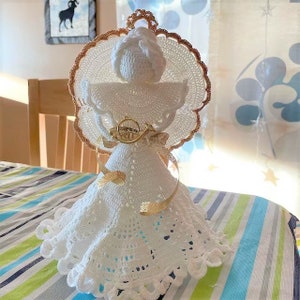 Puede incluir: Un adorno de &aacute;ngel de crochet blanco con un halo dorado y una cinta dorada. El &aacute;ngel lleva un vestido blanco con ribete de encaje y tiene un cuerno dorado.