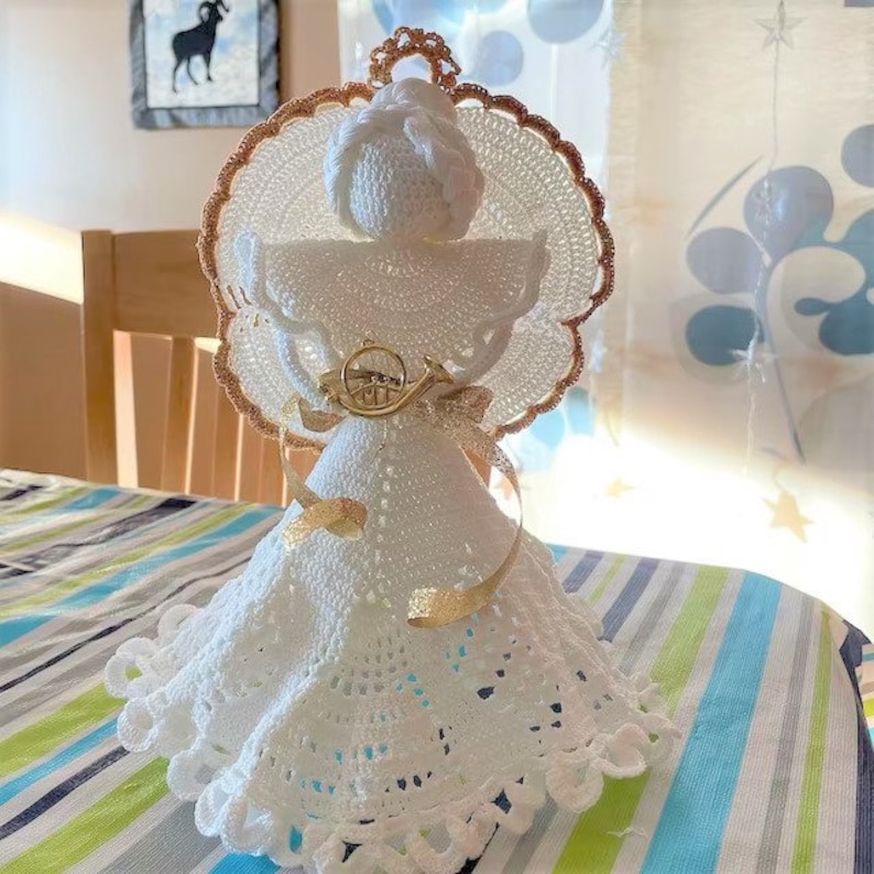 Puede incluir: Un adorno de &aacute;ngel de crochet blanco con un halo dorado y una cinta dorada. El &aacute;ngel lleva un vestido blanco con ribete de encaje y tiene un cuerno dorado.
