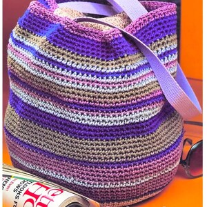 May include: A crochet bucket bag featuring horizontal stripes in shades of purple, lavender, and tan. The bag has a light purple strap and a patterned fabric lining visible at the top opening. The text PastPatternsPalooza is visible.