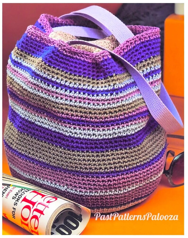 May include: A crochet bucket bag featuring horizontal stripes in shades of purple, lavender, and tan. The bag has a light purple strap and a patterned fabric lining visible at the top opening. The text PastPatternsPalooza is visible.