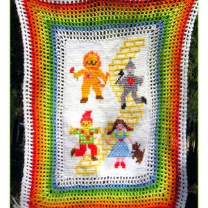 May include: A rectangular crochet blanket featuring a white center panel with a yellow brick road and four figures: a lion, a tin person, a scarecrow, and a person with a small dog. The border is a multi-colored rainbow pattern of red, orange, yellow, green, and blue.