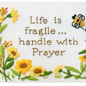 Puede incluir: Bordado con flores amarillas y hojas verdes que rodean el texto "Life is fragile... handle with Prayer". Una pequeña abeja bordada vuela sobre el texto.