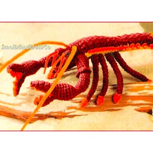 Puede incluir: Una langosta hecha a mano, de ganchillo, en tonos rojos, naranjas y amarillos. La langosta tiene pinzas, patas y antenas detalladas. El texto "SmallsandDollsCo" es visible. La langosta está sobre una superficie arenosa.