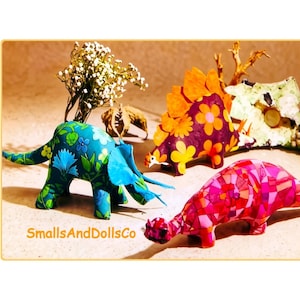 May include: Three dinosaur figurines featuring fabric patterns. One is teal with blue and green floral prints, one is purple with orange flowers and back plates, and one is pink with a geometric mosaic pattern. The text SmallsAndDollsCo is visible.