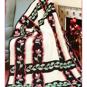 May include: A crocheted blanket with a floral design. The blanket features alternating stripes of cream and dark brown, with pink and green floral accents. The blanket is draped over a chair, showcasing its intricate pattern and texture.