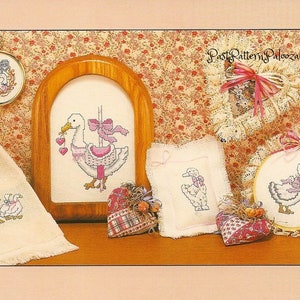 Pu&ograve; includere: Una collezione di ricami a punto croce con un'oca bianca con accenti rosa. I disegni sono esposti in varie cornici, tra cui un cerchio, un telaio quadrato e un telaio rettangolare. I disegni sono tutti su tessuto bianco e presentano una variet&agrave; di punti, tra cui il punto croce, il punto indietro e il punto raso.