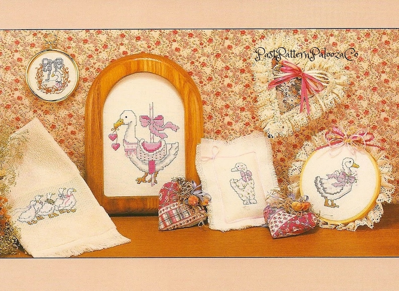 Pu&ograve; includere: Una collezione di ricami a punto croce con un'oca bianca con accenti rosa. I disegni sono esposti in varie cornici, tra cui un cerchio, un telaio quadrato e un telaio rettangolare. I disegni sono tutti su tessuto bianco e presentano una variet&agrave; di punti, tra cui il punto croce, il punto indietro e il punto raso.