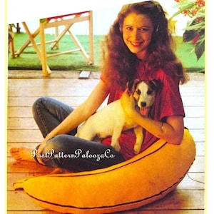 May include: A smiling person sits with a small dog on a large, yellow banana-shaped cushion. The person wears a red shirt and blue jeans. The cushion has a brown stem and the text "PastPatternPaloozaCo".