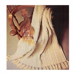 May include: A white crocheted blanket with a cable stitch pattern and a fringe border. The blanket is draped over a wooden chair.