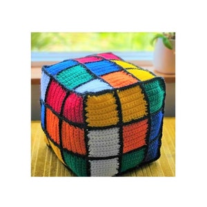 Pode incluir: Um travesseiro em forma de cubo de croch&ecirc; colorido com contornos pretos. O travesseiro &eacute; feito de quadrados de diferentes cores: vermelho, laranja, amarelo, verde, azul e branco.