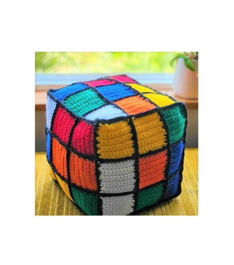 Pode incluir: Um travesseiro em forma de cubo de croch&ecirc; colorido com contornos pretos. O travesseiro &eacute; feito de quadrados de diferentes cores: vermelho, laranja, amarelo, verde, azul e branco.