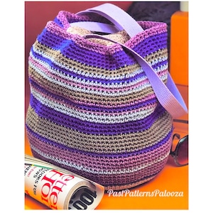May include: A crochet bucket bag featuring horizontal stripes in shades of purple, lavender, tan, and light gray. The bag has a wide, light purple fabric strap and a drawstring closure. A rolled magazine with visible text is positioned nearby.
