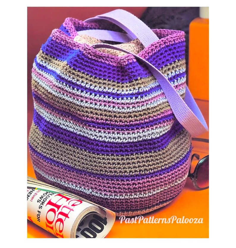May include: A crochet bucket bag featuring horizontal stripes in shades of purple, lavender, tan, and light gray. The bag has a wide, light purple fabric strap and a drawstring closure. A rolled magazine with visible text is positioned nearby.