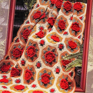 May include: A crochet blanket featuring a repeating pattern of hexagonal motifs in cream, orange, and dark green, accented with three-dimensional red rose appliqués. The textile is draped over a dark brown wooden frame, showcasing the intricate yarn work.