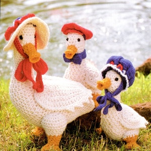 May include: Three crocheted goose figurines, each wearing a different colored hat. The largest goose is wearing a white hat with a red ribbon, the middle goose is wearing a red hat with a blue ribbon, and the smallest goose is wearing a blue hat with a blue ribbon.