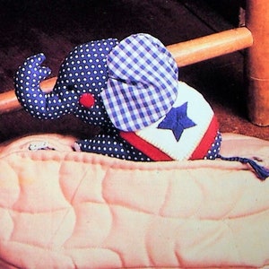 May include: A plush toy elephant with blue and white polka dots and a red heart on its face is peeking out of a beige peanut-shaped pillow. The elephant is wearing a blue and white gingham fabric on its head and a red, white, and blue star-shaped patch on its back.