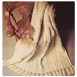 May include: An off-white knitted throw blanket featuring intricate cable knit patterns, raised bobble textures, and long fringe trim along the bottom edge. The text "Past Patterns Palooza" is printed in white script over the fabric surface.