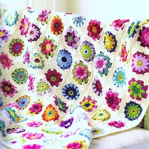 Peut inclure: Une couverture en crochet colorée avec un motif floral. La couverture est composée de carrés avec des fleurs de différentes couleurs au centre. La couverture est drapée sur un canapé blanc.
