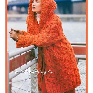 Puede incluir: Un cárdigan de punto naranja brillante con una capucha grande. El cárdigan tiene un patrón de punto de cable y se muestra siendo usado por una persona.