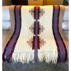 Peut inclure: Une couverture en crochet cr&egrave;me avec des franges et un motif g&eacute;om&eacute;trique. La couverture pr&eacute;sente un motif central de formes beiges, bordeaux et turquoise, encadr&eacute; de rayures verticales violettes et beiges. La frange est cr&egrave;me.