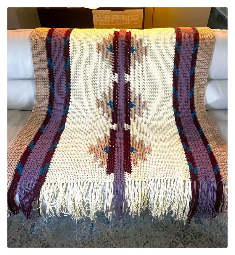 Peut inclure: Une couverture en crochet cr&egrave;me avec des franges et un motif g&eacute;om&eacute;trique. La couverture pr&eacute;sente un motif central de formes beiges, bordeaux et turquoise, encadr&eacute; de rayures verticales violettes et beiges. La frange est cr&egrave;me.