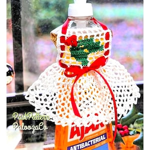 May include: A white crocheted cover for a dish soap bottle, featuring a red ribbon tie at the waist and a green and red holly leaf motif on the bodice. The cover is placed over an orange Ajax antibacterial dish soap bottle with white text.