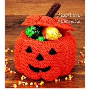 May include: A bright orange crocheted pumpkin container with a brown stem and a black stitched jack-o'-lantern face. The lid is partially open, revealing green and yellow wrapped candies inside. The text PastPattern PaloozaCo is visible.