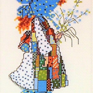 May include: Cross stitch pattern featuring a girl in a blue dress with a patchwork design, holding a bouquet of flowers. The pattern is titled "Holly Hobbie" and is from PastPatternPaloozaCo.