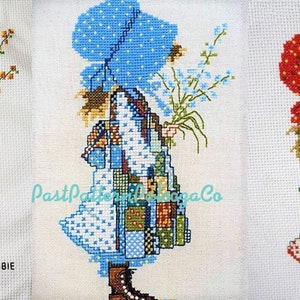 May include: Three cross-stitch patterns of a girl in a dress with a patchwork design, holding flowers. The girl is wearing a different colored bonnet in each pattern: brown, blue, and red.